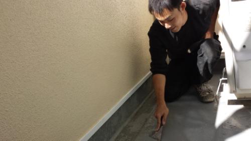 20130927-092947-2 | 外壁塗装は神奈川県横浜市の塗装職人 Man crouching beside a beige exterior wall, pointing at a seam along the base of the wall on a concrete surface near a metal strip.