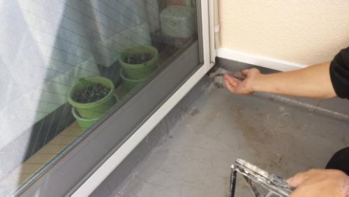 20130927-094423 | 外壁塗装は神奈川県横浜市の塗装職人 Person applying sealant along the edge of a balcony floor beside a sliding glass door, with green plant pots visible inside.