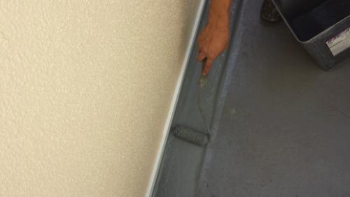 20130927-152442 | 外壁塗装は神奈川県横浜市の塗装職人 Close-up of a hand using a paint roller to apply gray paint along a seam between a beige wall and a gray floor surface.