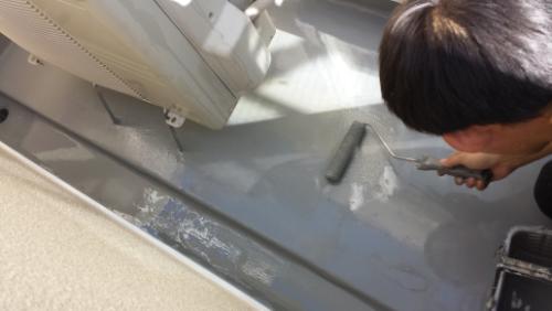 20130930-093927 | 外壁塗装は神奈川県横浜市の塗装職人 Person applying a gray floor coating with a hand trowel on a concrete floor near a wall, during construction work.