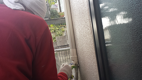 Person in a red jacket with a white hood and a glove, standing outside near an exterior wall and a vertical pipe or post, touching it with their hand.