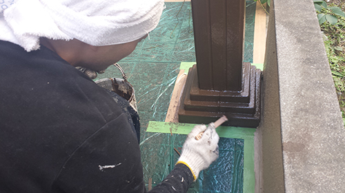 Person wearing a white head covering and gloves applies blue sealant around the base of a metal railing post outdoors.