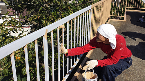 Person in a red shirt and white headscarf paints a white metal railing on an outdoor balcony, with a paint bucket nearby.