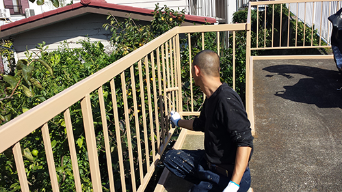 Person kneeling on an outdoor balcony, painting a beige metal railing with a brush, wearing gloves.