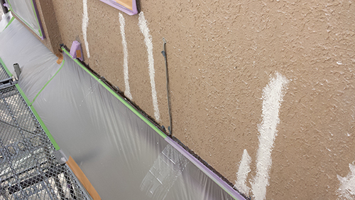 Exterior brown stucco wall under construction with protective plastic at the base, masking tape, and scaffolding nearby. White vertical sealant lines are visible on the wall.