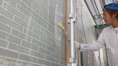 Worker in white shirt and blue cap rolls paint onto a gray brick wall from scaffolding.