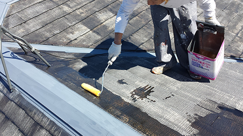 Person applying dark roof coating with a roller along a seam; paint can and open canister nearby, wearing gloves on a wooden roof surface.