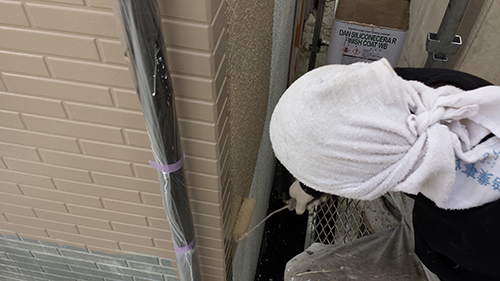 Maintenance worker with a white cloth over the head bending in a narrow outdoor space beside a beige wall and a metal pipe.