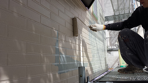 Painter wearing gloves rolls gray paint onto a brick wall from a scaffold platform outside a building
