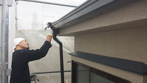 Maintenance worker wearing a white cap and gloves installs a gutter along the eaves of a beige house.