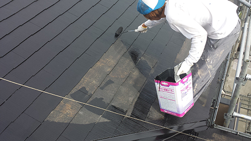 Worker in a white shirt and blue hard hat applies black roof coating with a roller on a sloped roof, using a pink-and-white paint can.