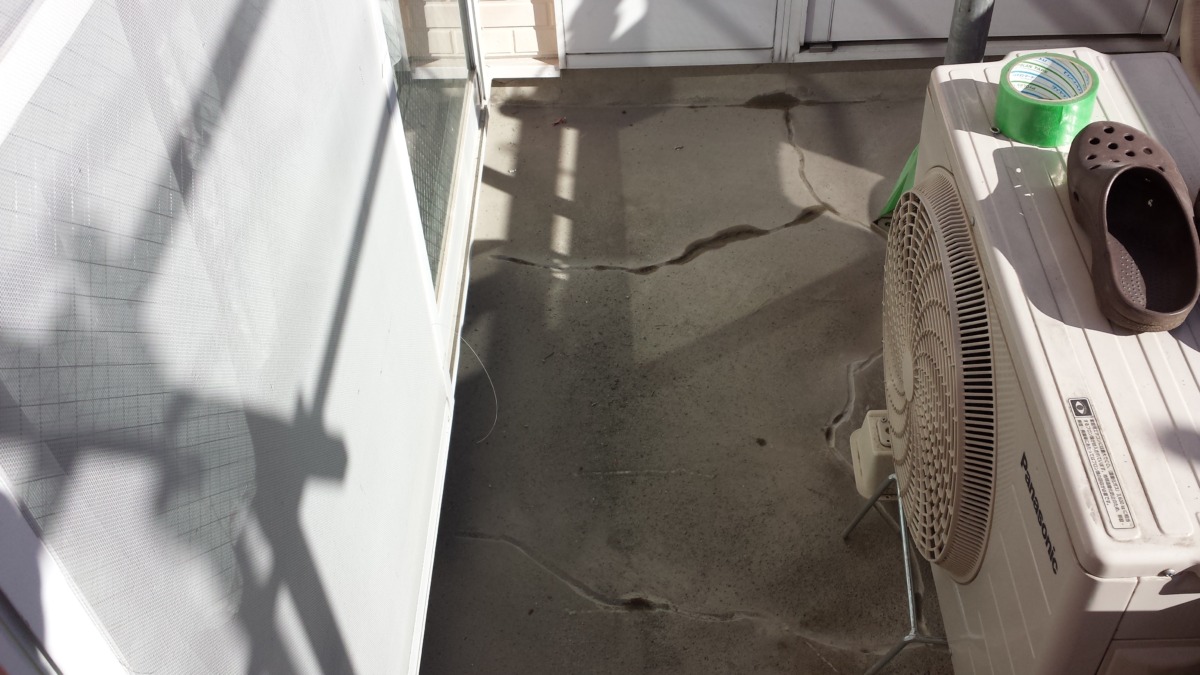 METADATA-START | 外壁塗装は神奈川県横浜市の塗装職人 Outdoor air conditioner on a concrete balcony with a brown clog resting on top and a roll of green tape nearby, sun casting shadows on the floor.
