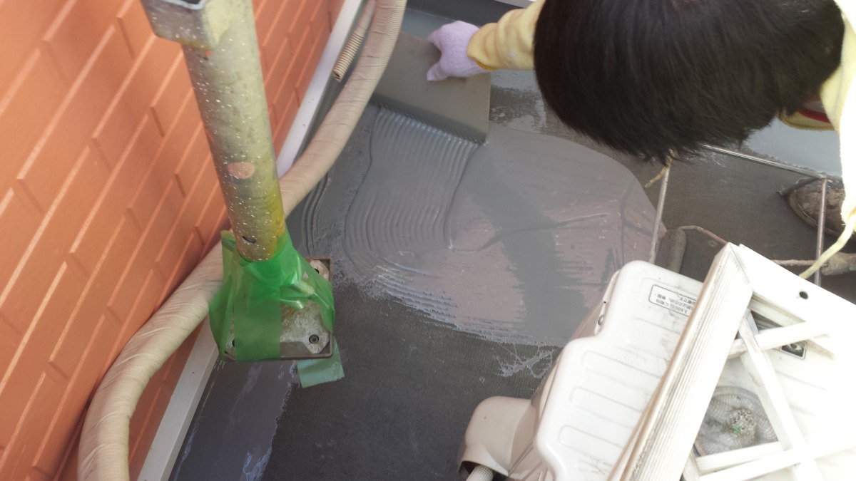 METADATA-START | 外壁塗装は神奈川県横浜市の塗装職人 Worker in yellow gloves troweling gray waterproof coating on a small balcony floor beside a brick wall.