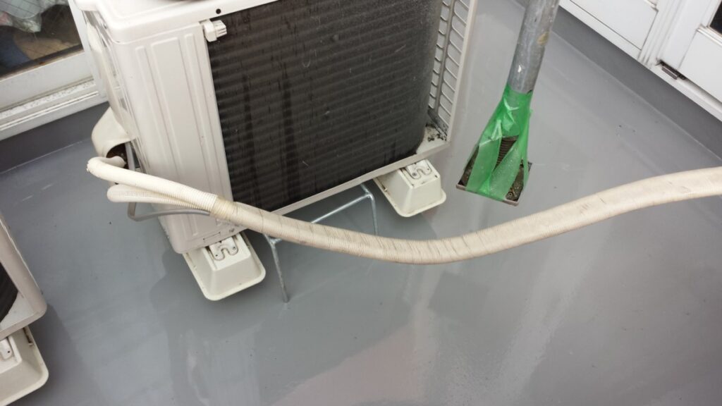 METADATA-START | 外壁塗装は神奈川県横浜市の塗装職人 Outdoor air conditioner condenser on a flat roof with insulated hoses and green taped pipe nearby, resting on white feet brackets.