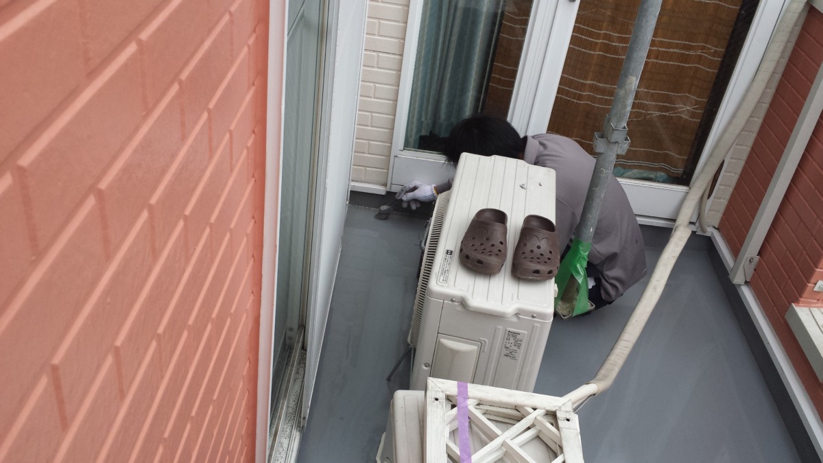 METADATA-START | 外壁塗装は神奈川県横浜市の塗装職人 Person crouching on a balcony next to an air conditioner, wearing gloves and brown clogs, performing maintenance outside a sliding door.