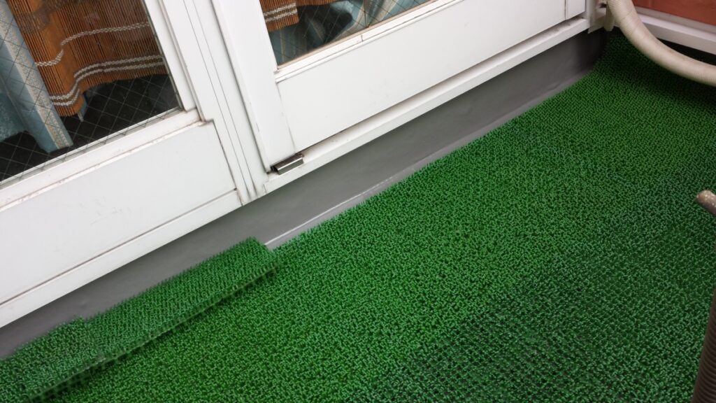 METADATA-START | 外壁塗装は神奈川県横浜市の塗装職人 Green artificial-grass outdoor mat laid along a glass door threshold with a rolled edge piece nearby