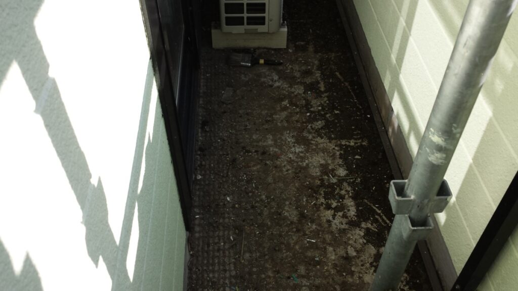 METADATA-START | 外壁塗装は神奈川県横浜市の塗装職人 Narrow outdoor utility space with a dirty concrete floor, an air conditioner unit at the far end, a bottle on the ground, and a metal railing on the right.