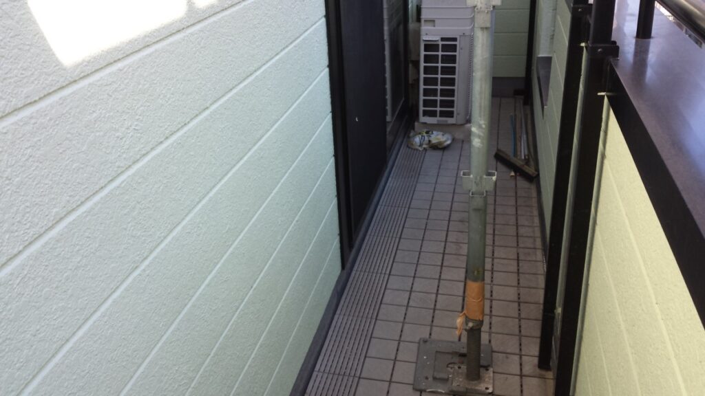 METADATA-START | 外壁塗装は神奈川県横浜市の塗装職人 Narrow outdoor alley between buildings with a vertical scaffolding pole taped at the base, a grated vent unit at the far end, and scattered debris on tiled floor.
