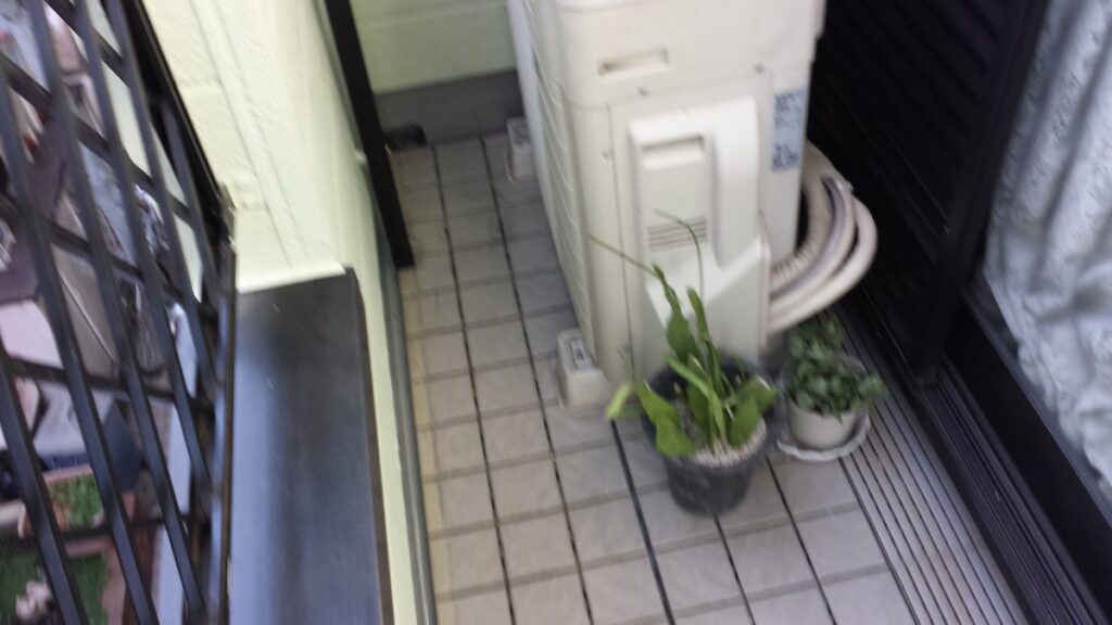 METADATA-START | 外壁塗装は神奈川県横浜市の塗装職人 Blurry balcony corner with a white air conditioner unit, two potted plants on a tiled floor, and coiled hoses beside the unit.