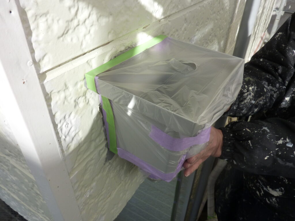 Worker wrapping an exterior light fixture in plastic with green and purple painter's tape to protect it during painting.