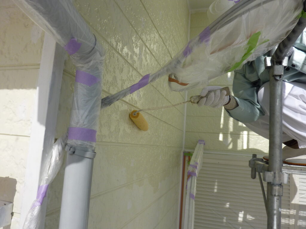 Worker on scaffolding painting an exterior wall with a roller, protected by plastic sheeting and purple painter's tape