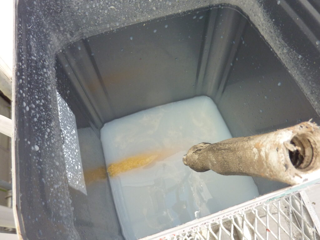 Open gray plastic bin with a rusty pipe pouring a yellowish liquid into a milky blue liquid at the bottom.