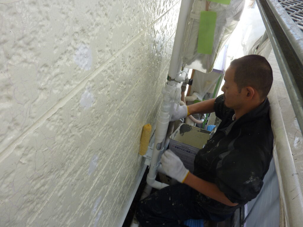 Worker in dark clothing wearing gloves handles white PVC plumbing pipes on an exterior wall.