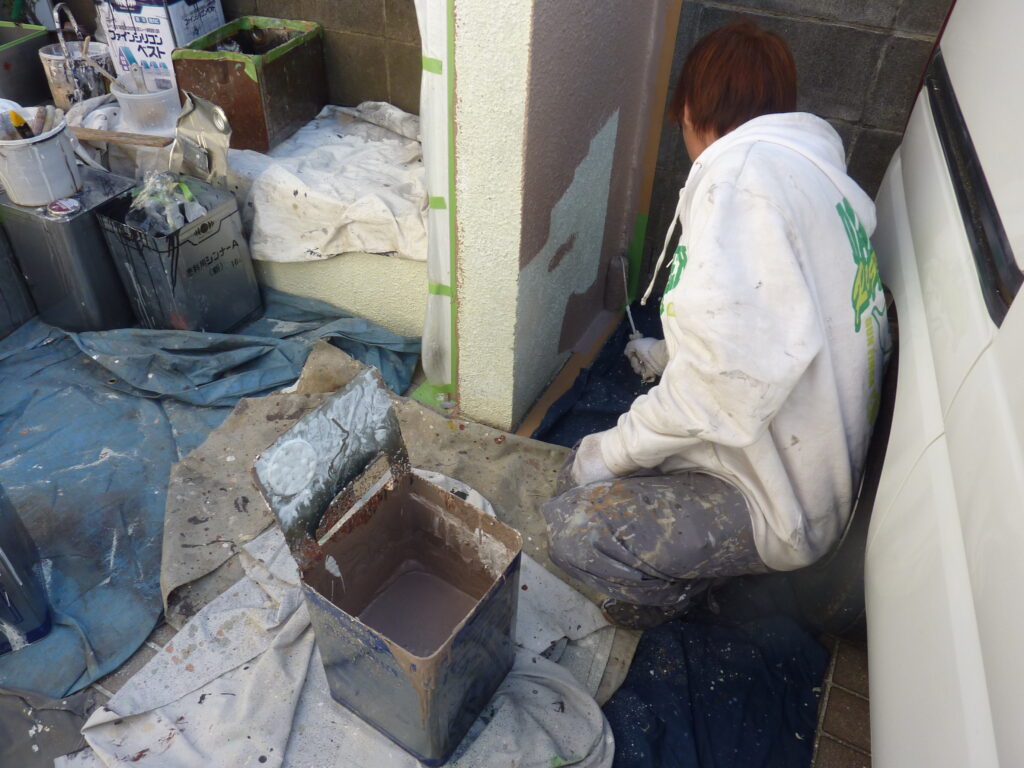 Person kneeling by a doorway, painting an exterior wall with a roller while wearing a white hoodie; construction supplies and paint cans around, with tarps on the ground.