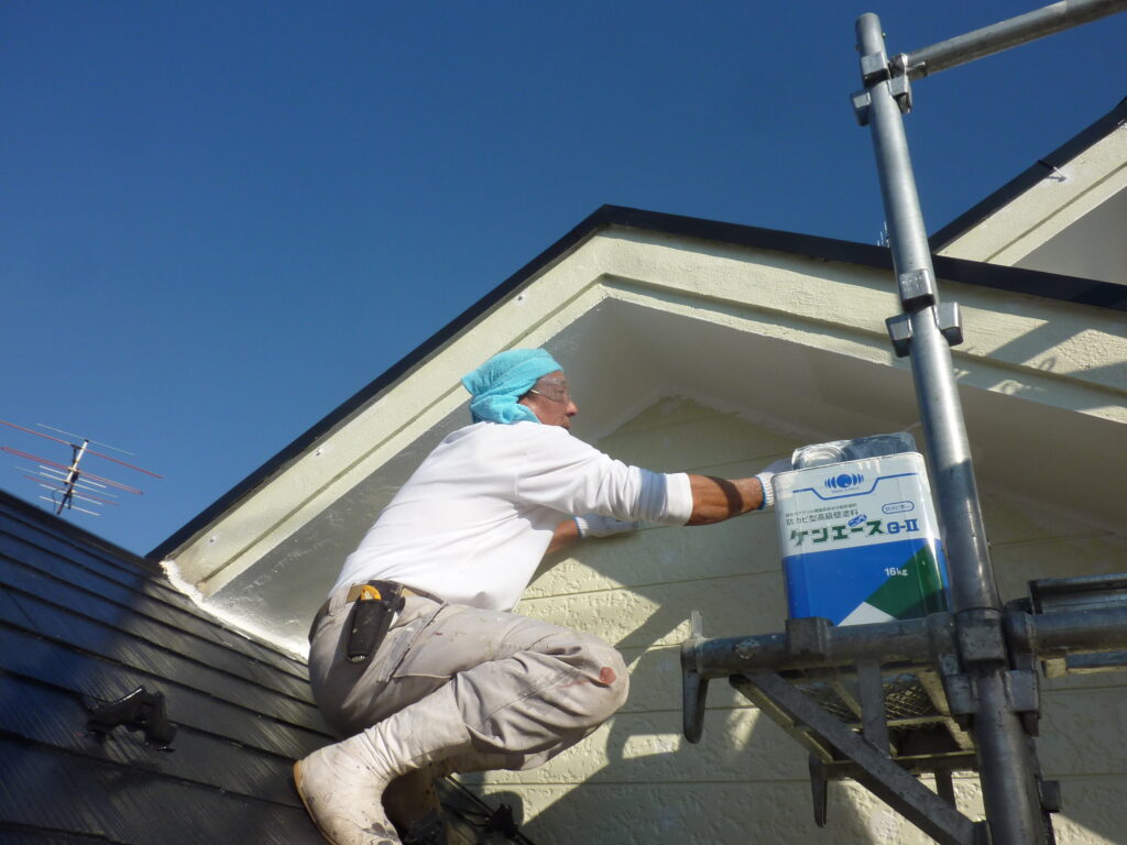 Construction worker kneels on a sloped roof, reaching toward a blue and white container on scaffolding under a clear blue sky.