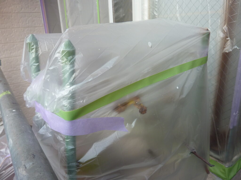 Outdoor appliance (likely an air conditioner) covered in clear plastic with masking tape to protect it during painting or renovation.