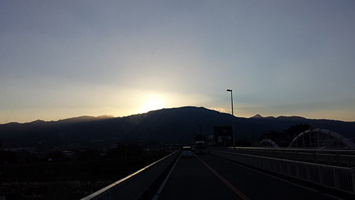 koushin_18yuuhi | 外壁塗装は神奈川県横浜市の塗装職人 Sun setting behind a mountain range as a road stretches toward the horizon on a bridge at dusk.