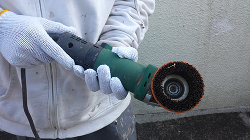 Person wearing gloves holds a green and black angle grinder with a grinding wheel, standing near a concrete wall.