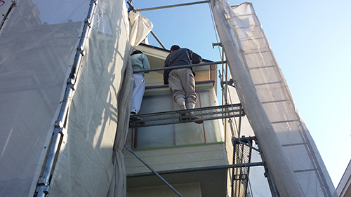 Two workers stand on metal scaffolding around a light-colored building, performing exterior renovation work.