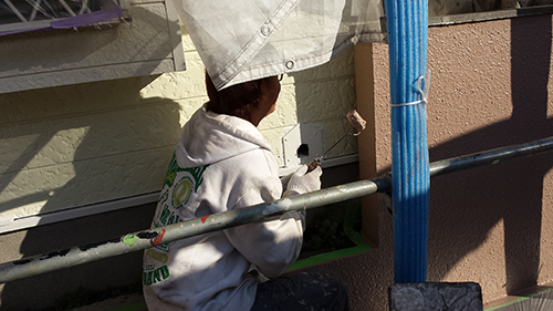 Person in a gray hoodie crouches on scaffolding, working on a small green planter box mounted to an exterior wall near a blue pole.