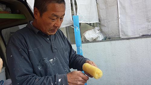 Man in a dark blue shirt painting with a yellow foam roller outdoors, near a vehicle and makeshift tarp area, focusing on the task.