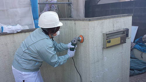 Construction worker in a helmet and mask using an orange-angle grinder to smooth a concrete wall beside a wall-mounted mailbox.
