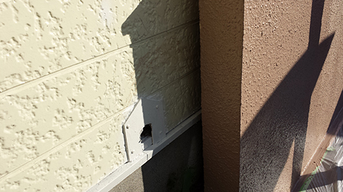 Small white wall vent with a rectangular opening mounted on a pale yellow textured exterior wall beside a brown column; sunlight casts a shadow on the right.
