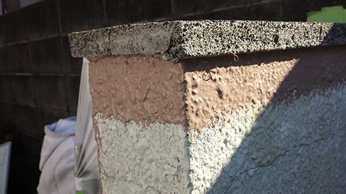 Close-up of a concrete corner with layered stucco in brown, beige, and pale green, with a rough cap and a shadowed edge.