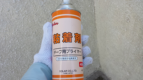Gloved hand holding a can of tape primer with Japanese text on an orange and white label, outdoor wall in the background.