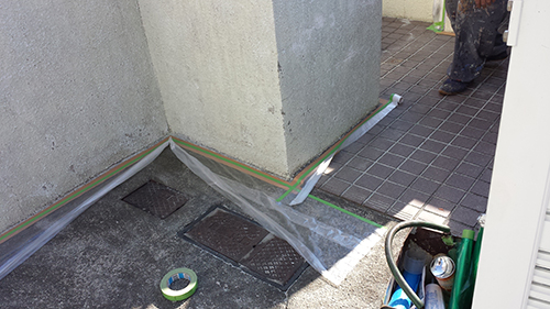 Corner of a building exterior with taped plastic sheeting and green painter's tape covering a sidewalk area, plus construction tools and hoses nearby.