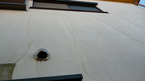 White stucco wall with a circular vent pipe near the bottom left and visible cracks; windows are at the top.