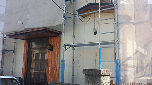Front view of a building under renovation with metal scaffolding, blue padding on the posts, and a brown wooden door on the left side.