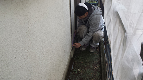 Person crouching on a narrow balcony, inspecting or sealing the base of the exterior wall with a tool.