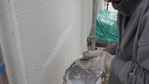 Worker in gloves applies plaster to a textured exterior wall, holding a bucket at a construction site.