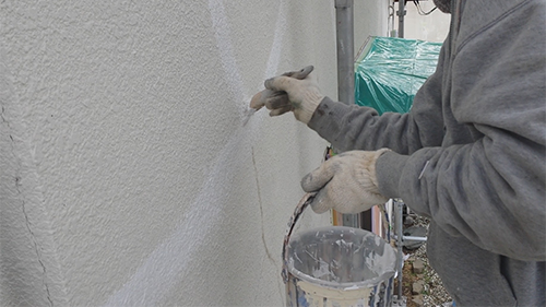 Worker in a gray sweatshirt applies plaster to an exterior wall using a trowel; a bucket of material sits nearby.