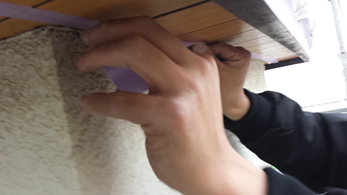 Close-up of hands sliding a purple edge trim under a wooden surface, installing it along a wall edge.