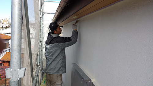 Construction worker on scaffolding applying white paint to an exterior wall under the eaves.