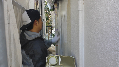 Person in a black-and-white cap and dark jacket rollers paint on a narrow outdoor wall.0 wall is textured and white, with a metal ladder visible in the background.