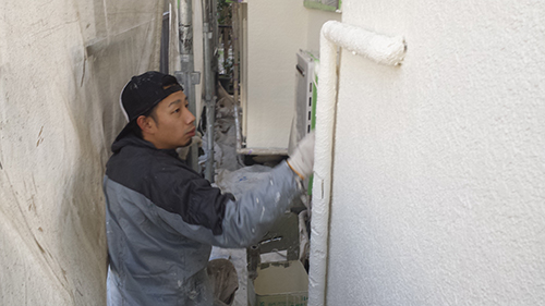 Man wearing a cap and jacket uses a paint roller to apply white paint to an exterior wall in a narrow alleyway outdoors