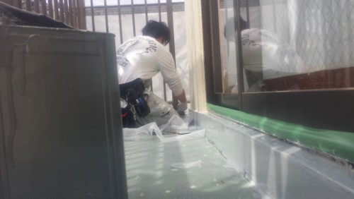 20140318-1138231 | 外壁塗装は神奈川県横浜市の塗装職人 Construction worker sealing a window frame on a flat exterior wall, kneeling with tools, plastic sheeting around the area